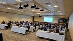 "Five rows of tables with white table clothes with people attentively looking forward at the speaker, who is not in frame, with a PowerPoint slide on the back wall describing solution architecture at the University of Manitoba