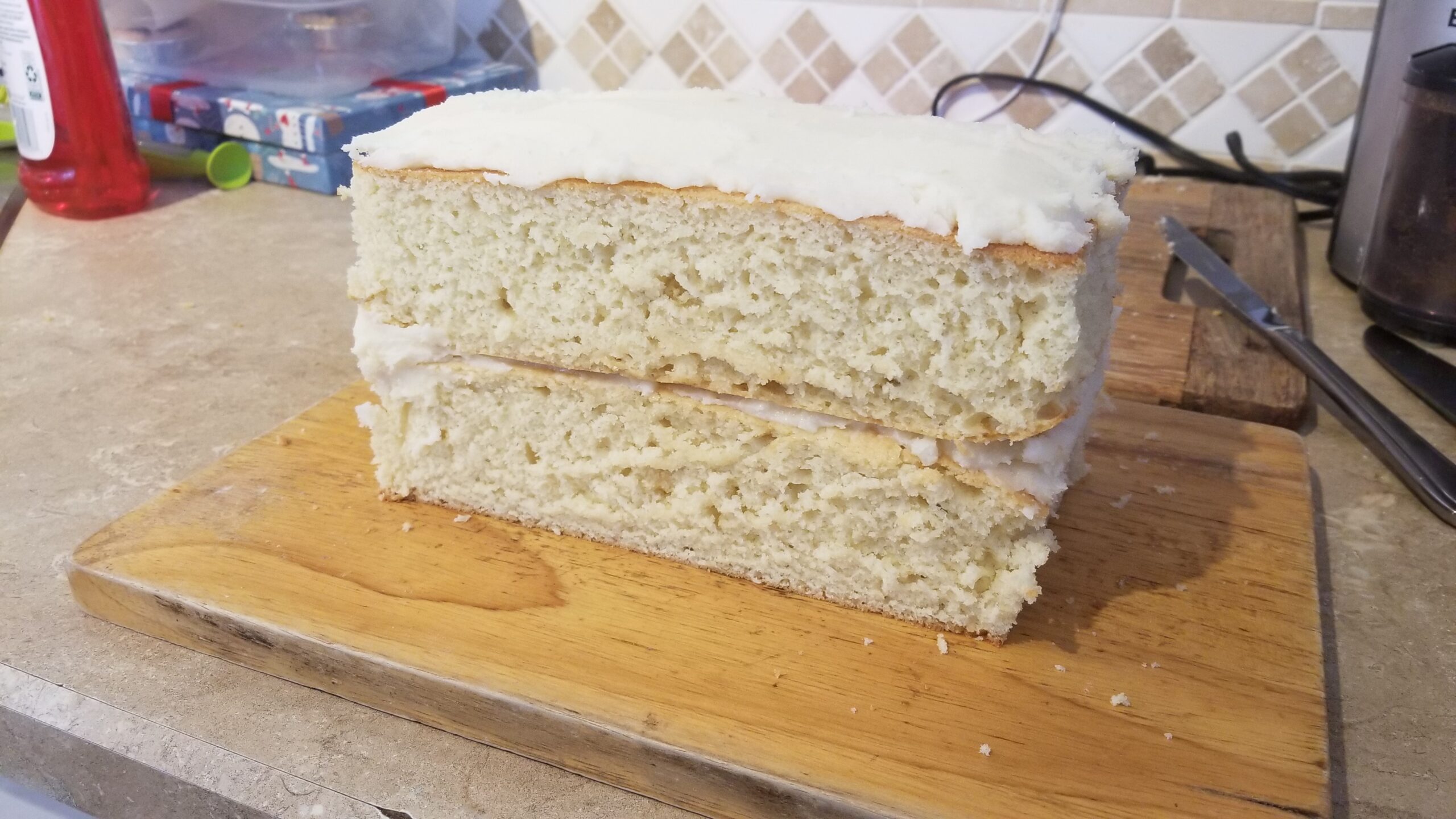 A picture of a homemade vanilla layer cake with white buttercream frosting between layers and on top, but not around the sides exposing the layers. The cake is resting on a wooden cutting board sitting on a kitchen counter.